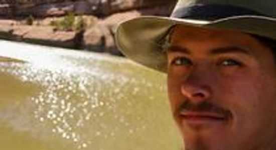 Man wearing a wide-brimmed hat by a sunlit river.