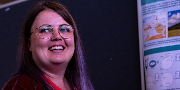 Smiling woman with glasses and purple hair in front of a presentation board.