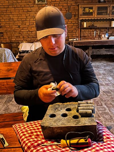Man wearing a cap sits at a table, handling small objects with a tool kit.