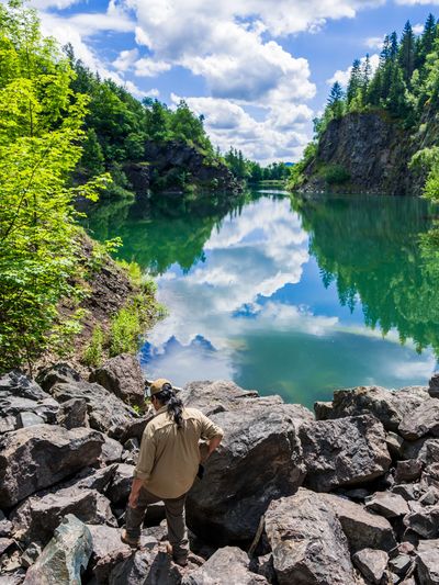 A person stands on rocks overlooking a serene lake surrounded by trees and cliffs.