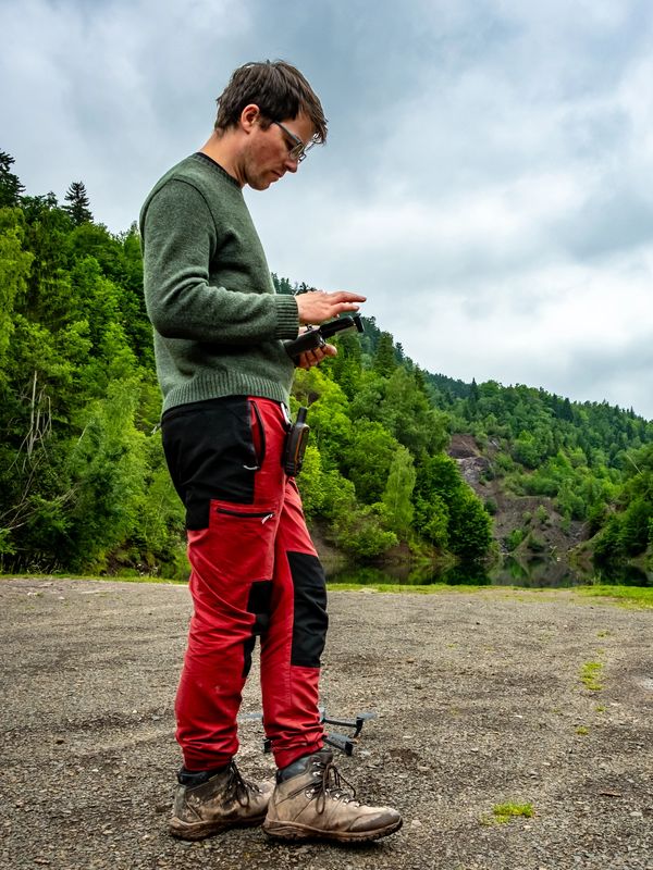 Man in red pants operating a drone controller in a forest clearing.