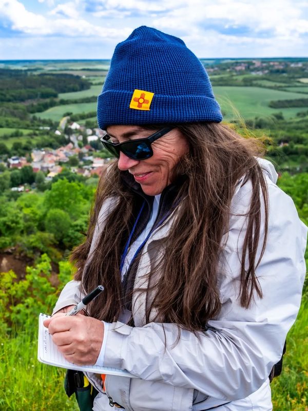 Woman in blue beanie and sunglasses writing in a notebook outdoors.