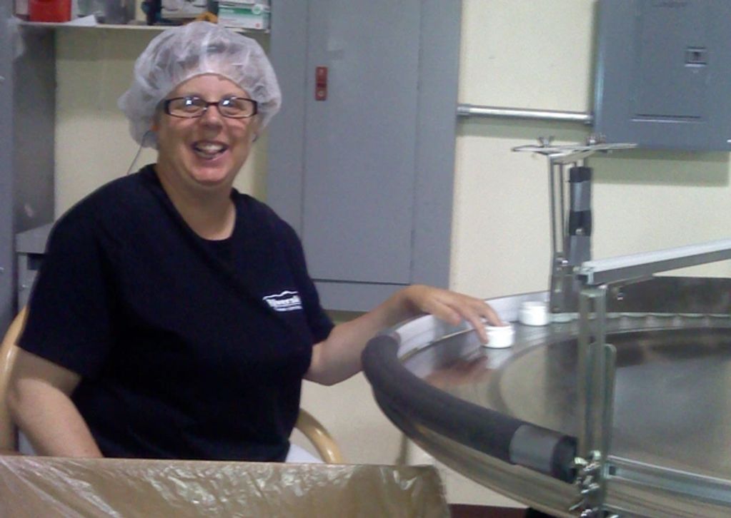 A smiling woman in a hair net loading jars onto a work table.
