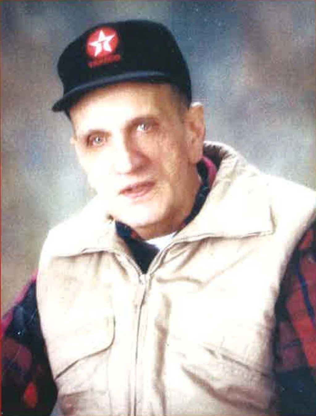 Portrait of older man wearing a black trucker hat and tan vest.