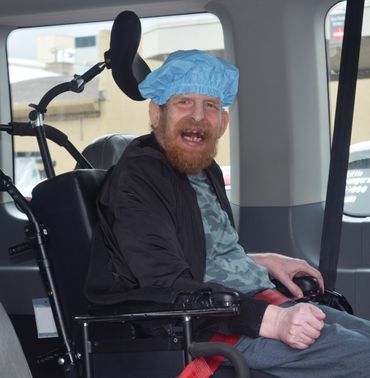 A smiling bearded man in a shower cap, seated inside a van in his wheelchair
