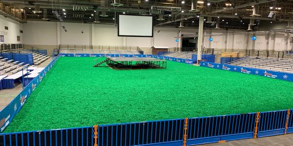 Indoor arena with green turf and bleachers surrounding a central platform.