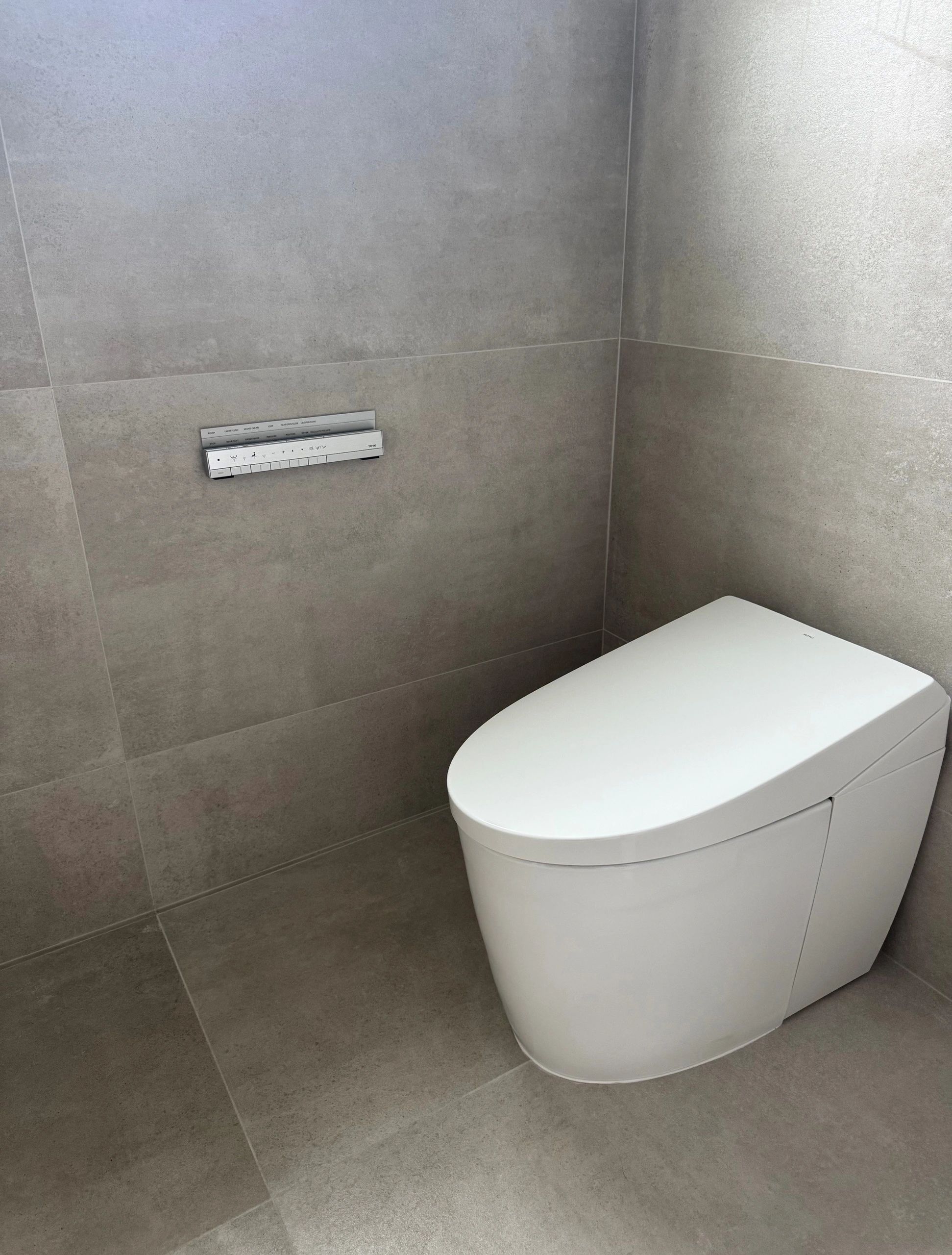 Modern white toilet with control panel on gray tiled wall.