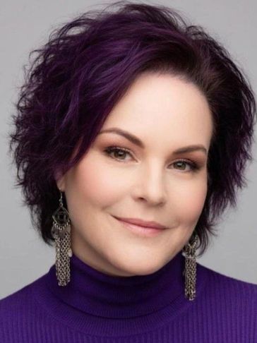 Woman with short purple hair and dangling earrings wearing a purple top.