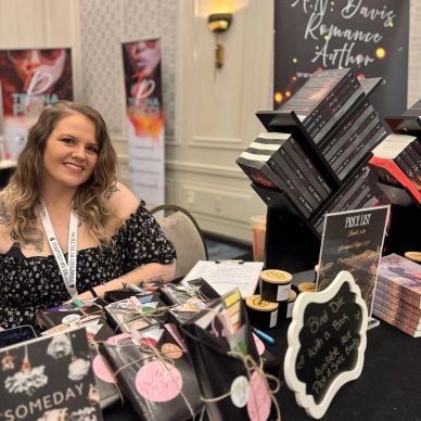 Smiling author at book signing table with books and promotional materials.