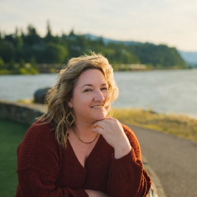 Woman smiling by a river during golden hour wearing a rust-colored sweater.