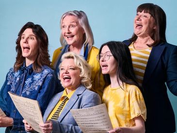 Five women happily singing together holding sheet music.