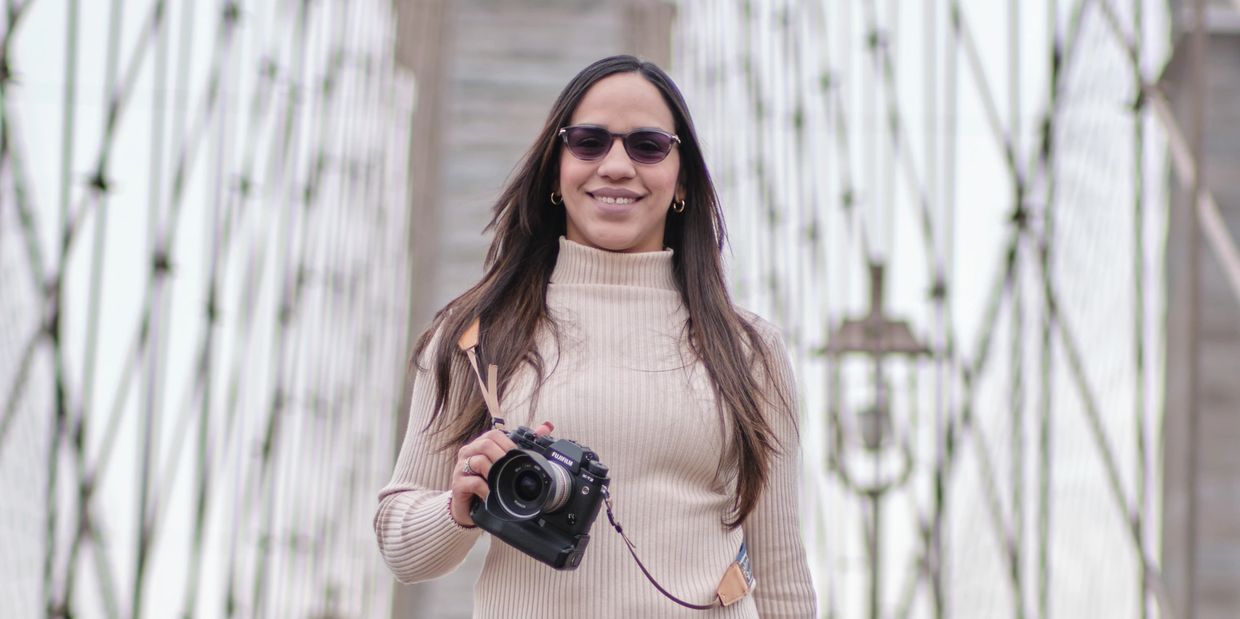 Woman with sunglasses holding a camera on a bridge, smiling.
