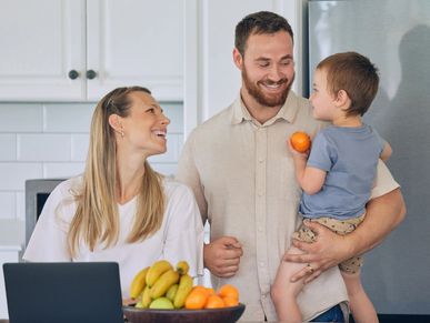 Young family in the kitchen with a laptop open and child in one parent's arms.