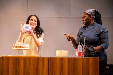 Two women interacting in a kitchen setting with a cake and a drink.
