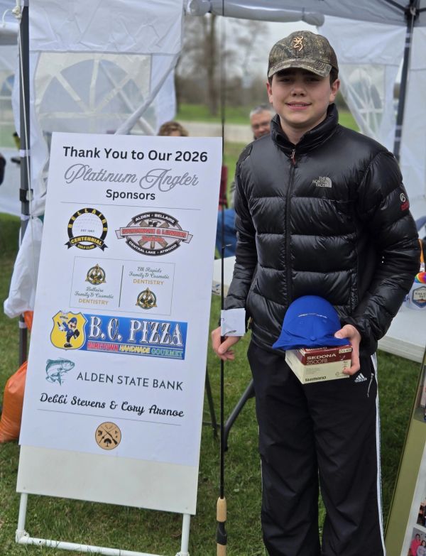 Young angler posing with fishing gear next to sponsor sign at outdoor event.
