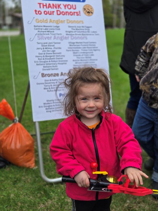 Smiling child in pink jacket holding a toy crossbow at an outdoor event.