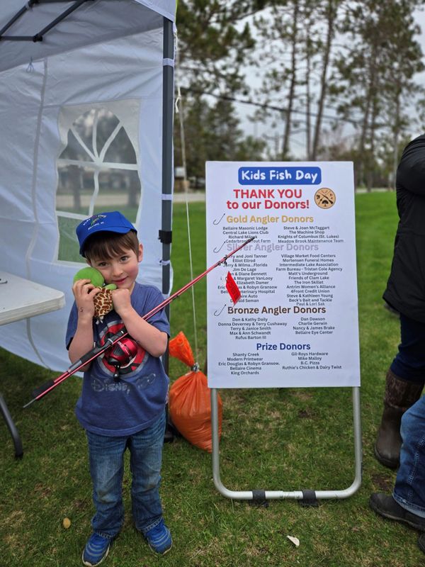 Young boy holding a fishing rod and toy, standing next to a Kids Fish Day thank you sign.