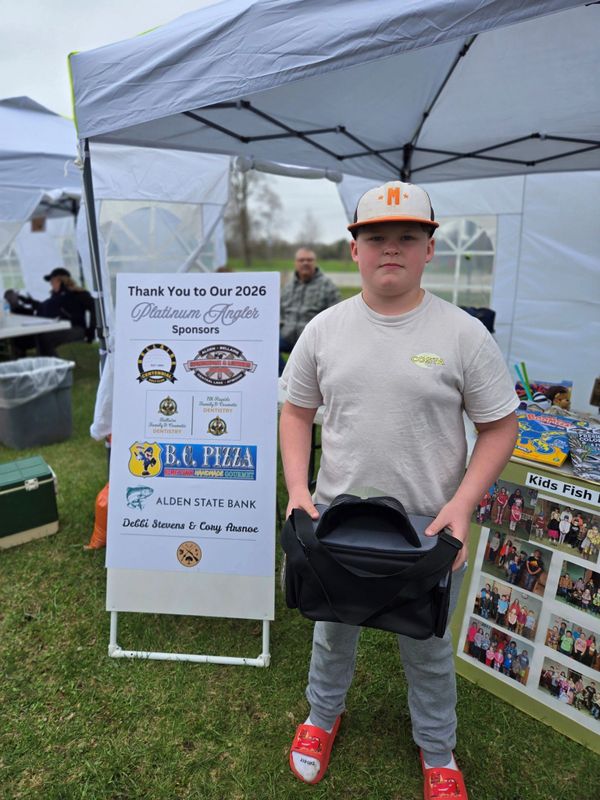 A boy stands at an outdoor event holding a black bag, next to a sponsor sign and photo display.