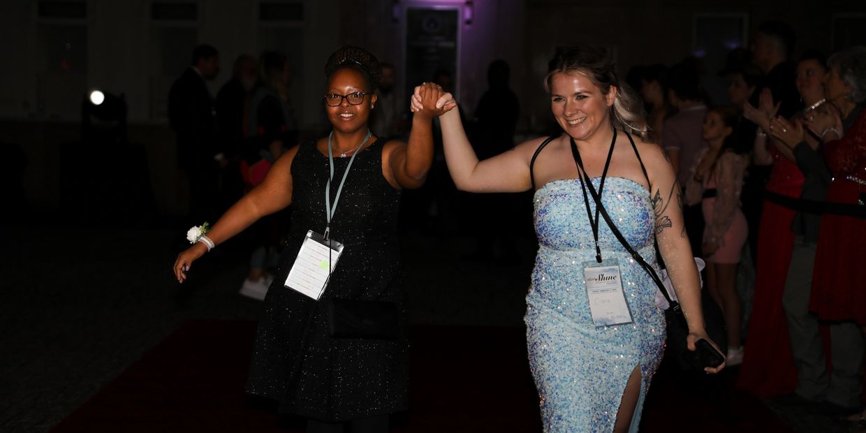 Two women holding hands and smiling while walking on a red carpet at night.