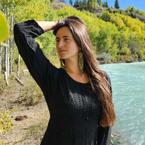 Woman in black and red stands by a river with green trees and blue sky.