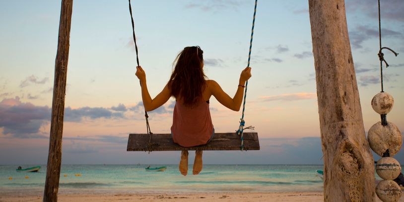Person swinging on a wooden swing facing the ocean at sunset.