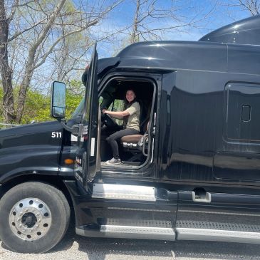 Person sitting in the driver's seat of a black semi-truck with the door open.