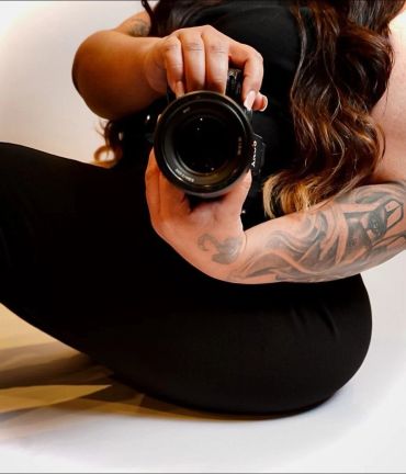 Person with tattoo holding a camera, sitting on the floor.