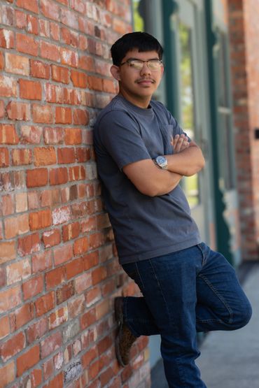 Young man leaning against a brick wall with arms crossed, wearing glasses and casual clothes.