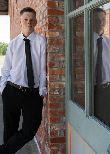 Young man in formal attire leaning against a brick wall with hands in pockets.