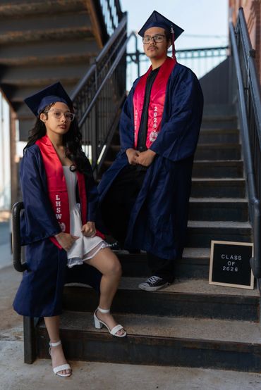 Two graduates in navy gowns and red stoles pose on stairs with a Class of 2026 sign.