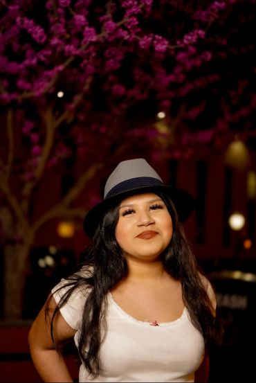 Young woman smiling, wearing a hat, with a flowering tree in the background.