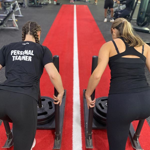 Two women pushing weighted sleds in a gym on a red track.