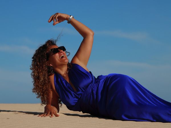 Woman in blue dress joyfully lounging on sand under clear sky.
