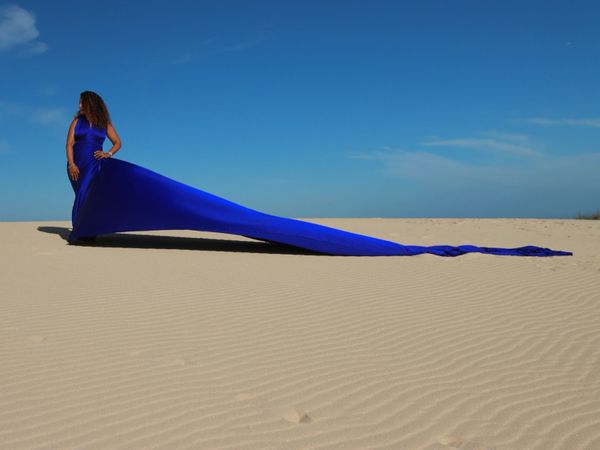 Woman in a flowing blue dress stands in sandy desert under clear sky.