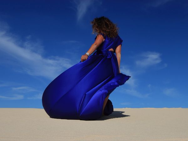 Woman in flowing blue dress stands on sand under a bright blue sky.