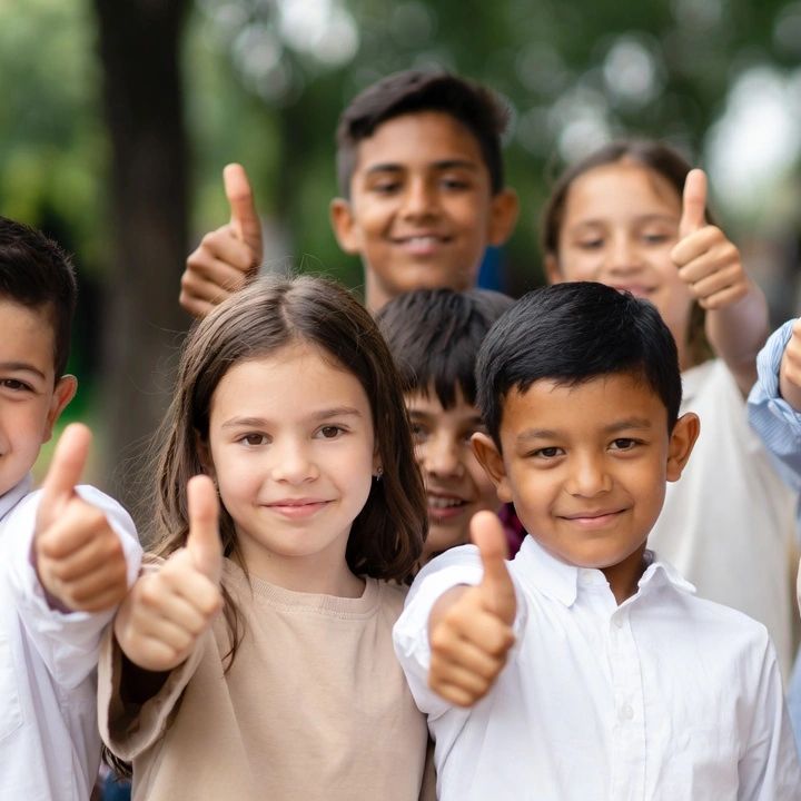A diverse group of children smiling and giving thumbs up outdoors.