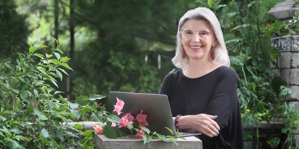 Smiling elderly woman with glasses using a laptop outdoors surrounded by greenery and flowers.