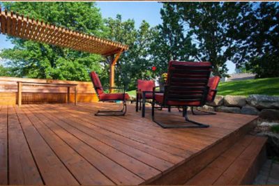 A wooden deck with outdoor chairs and a pergola in a green backyard.