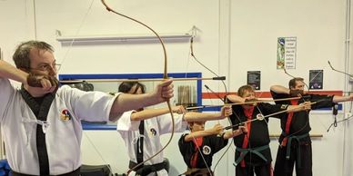 Group practicing archery in martial arts uniforms indoors.
