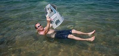 Tourist floating in the Dead Sea, Israel, reading a newspaper on a Dead Sea, Masada, and Qumran tour.