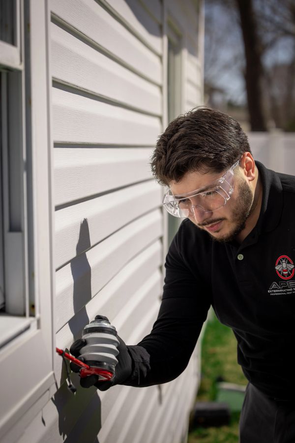 Man in safety goggles applying pest control treatment on house exterior.