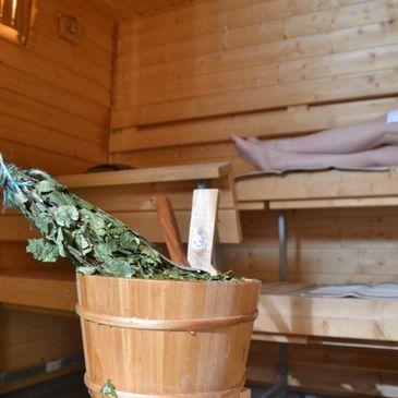 A wooden bucket with leaves inside in a sauna with someone relaxing.