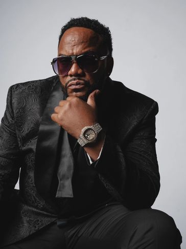 Man in a black patterned suit and sunglasses with a sparkling watch poses thoughtfully.