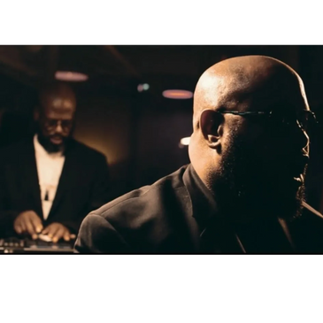 Two men in a dimly lit room, one focused on a device, the other in shadow.