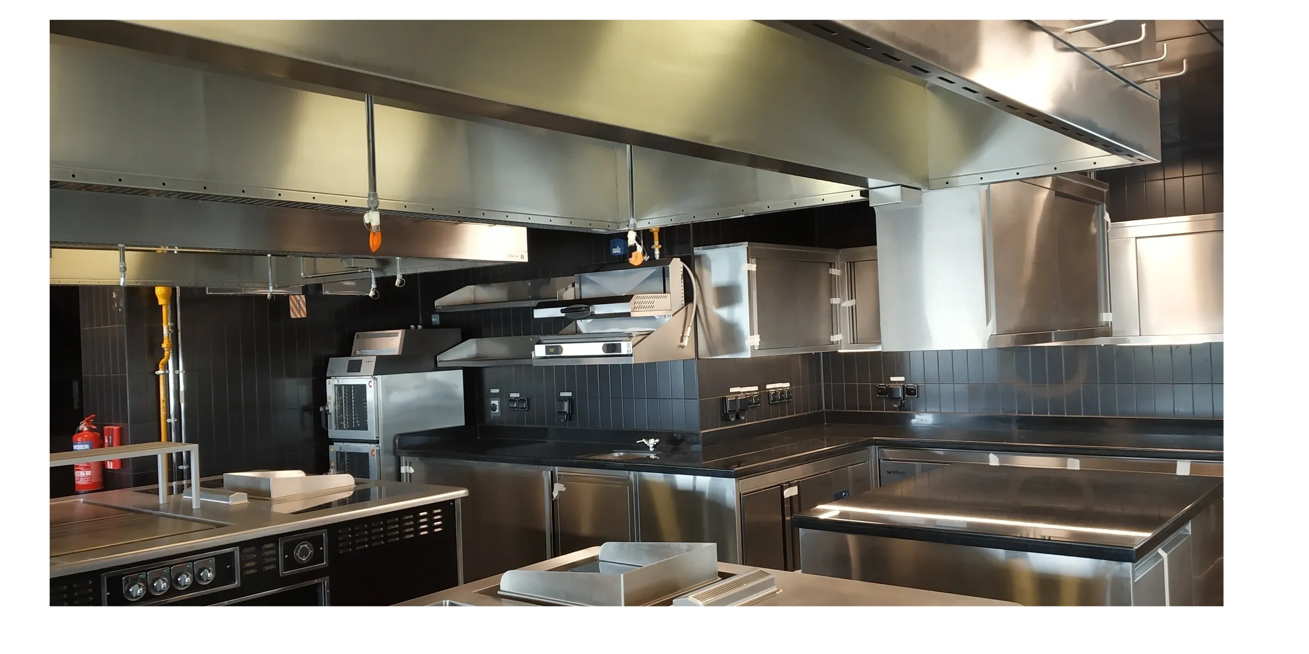 Modern commercial kitchen with stainless steel appliances and black tiled walls.