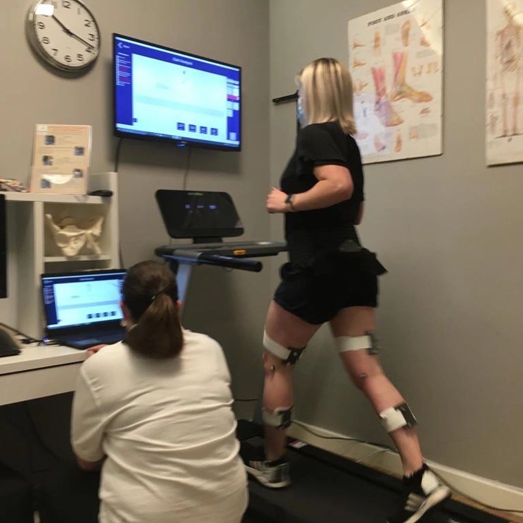 Woman running on treadmill with sensors, monitored by a professional in a clinical setting.