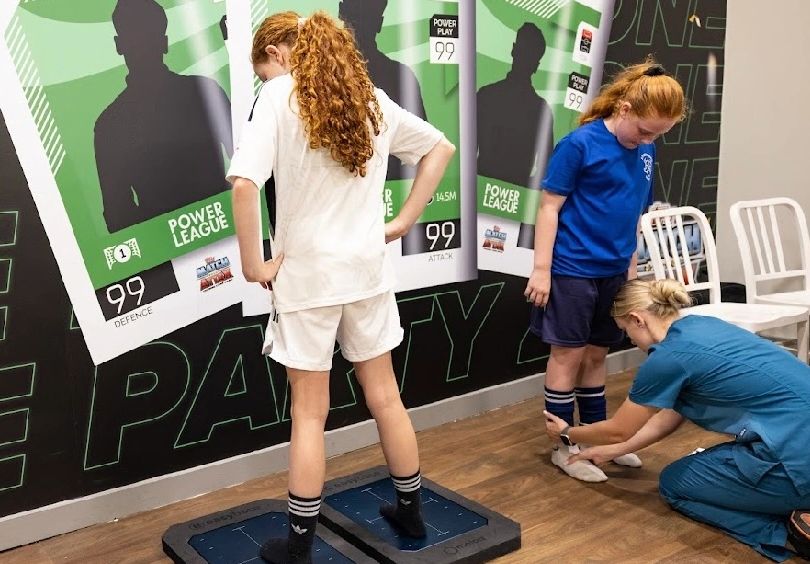 A sports therapist fitting a soccer shoe on a girl while another girl stands on a platform.