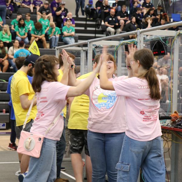 Three women in pink shirts celebrate with high-fives at a robotics event.