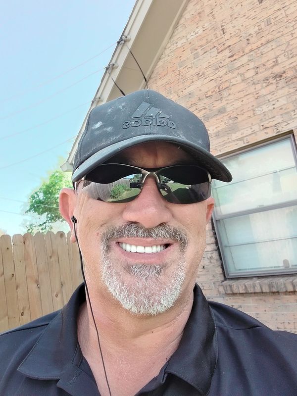 Man wearing sunglasses and a black Adidas cap smiles outdoors.