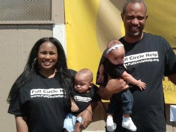 A smiling family wearing matching Full Circle Help t-shirts holds two babies outdoors.